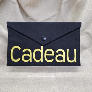 Op de afbeelding: Zwarte envelop-achtige etui met gele tekst "Cadeau". De etui heeft een klepsluiting met een enkele zwarte knoop. De achtergrond is een neutrale beige stof.