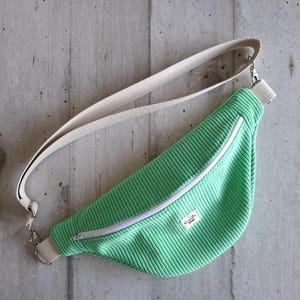 Peut inclure: Sac banane en velours côtelé vert menthe avec fermeture éclair blanche et bandoulière réglable. Le sac comporte une étiquette rectangulaire avec du texte. La bandoulière est blanc cassé et fixée avec du matériel métallique argenté.