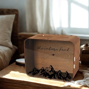 May include: A wooden money box with the words "Adventure Fund" and a mountain range design. The box has a slot on top for inserting money and a clear front panel. The design includes an airplane and dotted line.