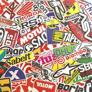 May include: A selection of vibrant stickers displaying logos and text from various brands. The stickers, in diverse shapes and sizes, include names such as Honda, Motul, and Brembo. Predominant colours are red, white, black, and yellow.