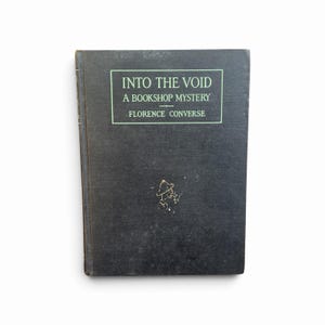 May include: A black hardcover book titled Into the Void: A Bookshop Mystery by Florence Converse. The front cover features a rectangular green border enclosing the title and author text, with a small, faded gold illustration of a person below.