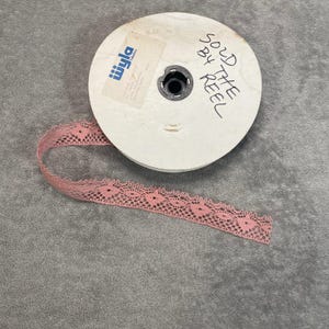May include: A spool of pink lace ribbon with an openwork pattern. The white spool has a label and the text "SOLD BY THE REEL". The ribbon is displayed on a grey surface. The lace is a soft pink colour.