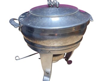 Pode incluir: Um prato de serviço de metal prateado com uma tampa redonda que apresenta uma borda de contas e uma alça em forma de laço. O recipiente tem duas alças laterais com textura martelada e repousa sobre um suporte de três pernas com queimador.