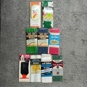 Könnte beinhalten: Eine Sammlung von Nähzubehör, darunter Schrägband, Nahtband und Rick Rack. Die Verpackungen sind in verschiedenen Farben wie Pfirsich, Grün, Blau, Rosa, Weiß und Schwarz erhältlich. Zu den Marken gehören Wrights und Trimtex.