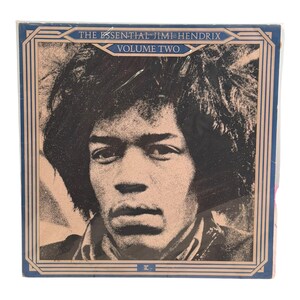 May include: A square vinyl record cover featuring a high-contrast, sepia-toned portrait of Jimi Hendrix. The text reads THE ESSENTIAL JIMI HENDRIX VOLUME TWO. The cover is framed by a dark blue border with geometric lines and a small logo at the bottom.
