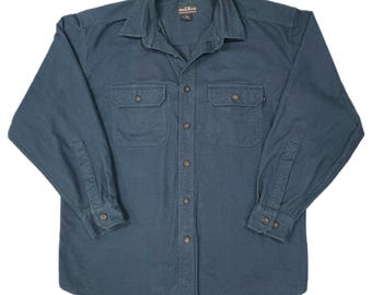 May include: A long-sleeved, teal-colored button-down shirt with two chest pockets. The shirt has a collared neckline and buttoned cuffs. The brand name "Woolrich" is visible on the collar.