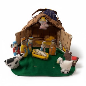 May include: A soft fabric nativity set featuring a brown stable with a blue roof and a hanging angel ornament labelled Peace on Earth. The set includes plush figures of a baby in a manger, adults, a cow, a sheep, and a donkey on a green base.