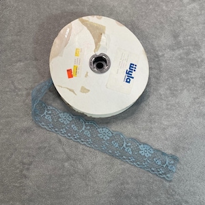 May include: A roll of pale blue lace with a floral design. The lace is partially unrolled, showing its intricate pattern. The roll is white with a metal core and a label.