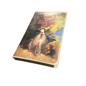 Op de afbeelding: Een vintage VHS-tape van "The Lord of the Rings" met een fantasie-illustratie op de cover. De illustratie toont een tovenaar met een gloeiend zwaard, met twee kleine figuren aan de onderkant. De titel staat in rode letters.