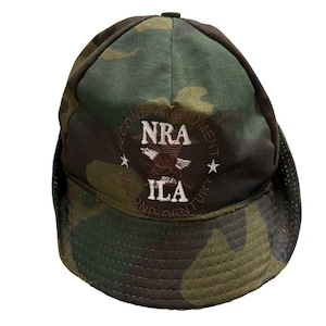 Puede incluir: Una gorra de béisbol de camuflaje con un logotipo bordado en la parte delantera. El logotipo muestra un gráfico de águila con el texto NRA e ILA, rodeado por el texto circular SECOND AMENDMENT SECOND CENTURY y dos estrellas blancas a los lados.