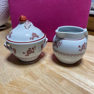 Pode incluir: Uma açucareira de cerâmica branca com tampa e um pequeno cremeira. Ambas as peças são decoradas com um padrão floral e acabamentos vermelhos. A açucareira tem duas pegas e um botão vermelho na tampa. A cremeira tem uma pega e um bico.