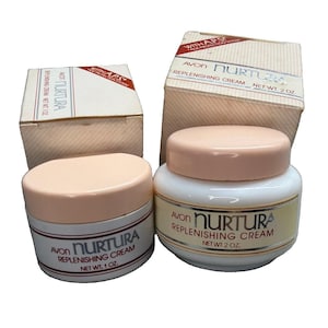 May include: Two jars of Avon Nurtura Replenishing Cream with matching boxes. One jar is 1 oz and the other is 2 oz. Both jars are white with peach-coloured lids and feature the text Avon Nurtura Replenishing Cream with APS Age Protection System.