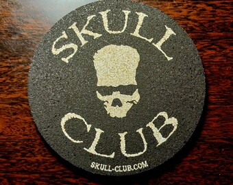 May include: A round, dark gray coaster with the words "SKULL CLUB" in a light beige font. A skull graphic is centered between the words. The website "SKULL-CLUB.com" is printed at the bottom.