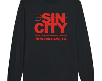 May include: Black long-sleeve shirt with the red text "SIN CITY" and "JUST THIS SIDE OF BAT COUNTRY NEW ORLEANS, LA". The shirt has a crew neck and long sleeves. The text is in a bold, sans-serif font.