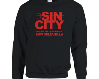 May include: Black sweatshirt with the red text "SIN CITY" and the phrase "JUST THIS SIDE OF BAT COUNTRY NEW ORLEANS, LA". The sweatshirt is a Gildan brand.