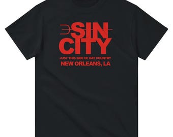 May include: Black t-shirt with the red text "SIN CITY" and the phrase "JUST THIS SIDE OF BAT COUNTRY NEW ORLEANS, LA". The shirt is a short-sleeved crew neck.