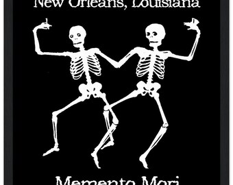 May include: Black framed art print featuring two white dancing skeletons. The text "New Orleans, Louisiana" is at the top, and "Memento Mori" is at the bottom. The skeletons are holding up their hands in a celebratory pose.