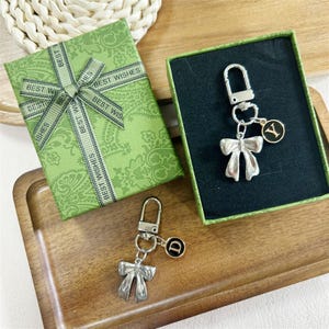 Peut inclure: Deux porte-clés argentés en forme de nœud papillon, l'un avec un charme en forme de lettre "Y" et l'autre avec un "D". Les porte-clés sont présentés dans une boîte verte et sur un plateau en bois, à côté d'une boîte cadeau verte avec un ruban "Best Wishes".