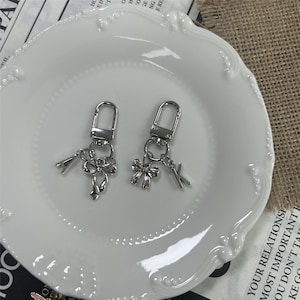 Peut inclure: Deux porte-clés argentés avec fermoirs mousquetons. Chaque porte-clés comporte un charm en forme de nœud et un charm en forme de lettre, l'un avec la lettre "A" et l'autre avec la lettre "K". Les porte-clés sont présentés sur une assiette blanche.