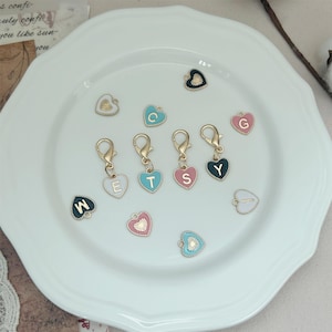 Può includere: Una collezione di ciondoli a forma di cuore in vari colori, tra cui nero, rosa, blu e bianco, con chiusure dorate. Ogni ciondolo presenta una lettera, come "E", "T", "S" e "Y". I ciondoli sono disposti su un piatto bianco.