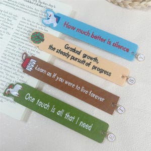 Peut inclure: Quatre marque-pages en feutre colorés avec des citations inspirantes. Les marque-pages sont bleus, beiges, marron et verts, chacun avec du texte brodé en blanc. Chaque marque-page a un petit charme avec une lettre.