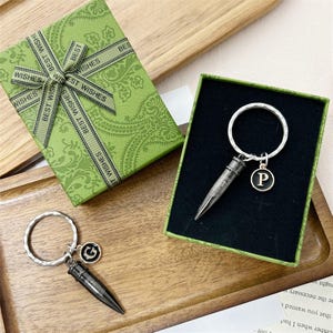 Puede incluir: Llaveros con un dije en forma de bala y un llavero circular. Un llavero tiene un colgante con la letra "P", el otro tiene un colgante "G". Los llaveros se exhiben en una caja de regalo verde con una cinta "Best Wishes".