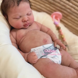 Könnte beinhalten: Eine lebensechte Babypuppe mit geschlossenen Augen, die eine Windel mit dem Text "Crissoso Full silicone doll" trägt. Die Puppe liegt auf einem cremefarbenen Kissen in einem geflochtenen Korb. Ein Schnuller ist sichtbar.