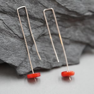 Pode incluir: Um par de brincos de metal prateado com um design de fio longo, fino e em forma de U. Cada brinco é finalizado com um pequeno disco redondo vermelho brilhante na extremidade inferior do fio, criando uma estética linear e pendente.