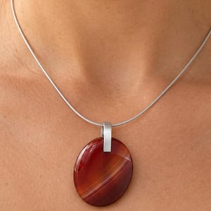 Könnte beinhalten: Eine Halskette mit einem glatten, ovalen Anhänger aus rot-orange gebändertem Achat, der an einem dünnen, silberfarbenen Metalldraht hängt. Der Anhänger ist oben mittig mit einer schlichten, matt silberfarbenen Metallöse befestigt.