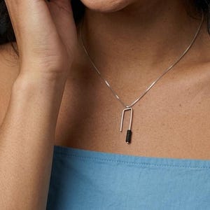 Könnte beinhalten: Eine silberne Halskette mit einem dünnen, U-förmigen Anhänger. Eine Seite des Anhängers ist aus glattem Silber, während die andere Seite mit einem strukturierten schwarzen Material umwickelt ist. Der Anhänger hängt an einer feinen Silberkette.