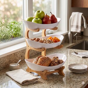 May include: A three-tiered serving stand with a light wood frame and three white, oval-shaped ceramic bowls. The top bowl holds green apples and red pears. The middle bowl contains walnuts and dried apricots. The bottom bowl holds bread.