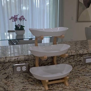 May include: A three-tiered serving stand featuring three white, oval-shaped ceramic bowls stacked vertically. The bowls are supported by a light-colored wooden frame with curved uprights and horizontal crossbars connecting each level securely.