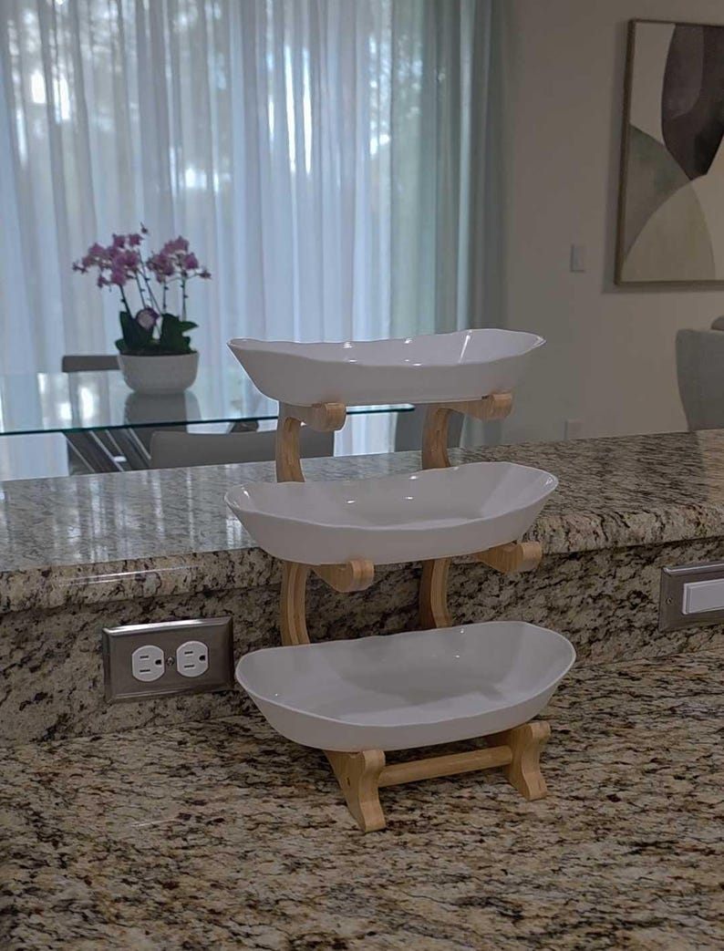 May include: A three-tiered serving stand featuring three white, oval-shaped ceramic bowls stacked vertically. The bowls are supported by a light-colored wooden frame with curved uprights and horizontal crossbars connecting each level securely.