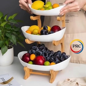 May include: A three-tiered fruit stand featuring white ceramic bowls supported by a light-colored wooden frame. The bowls are filled with assorted fresh fruit, including red apples, dark purple grapes, yellow lemons, and small orange citrus fruits.