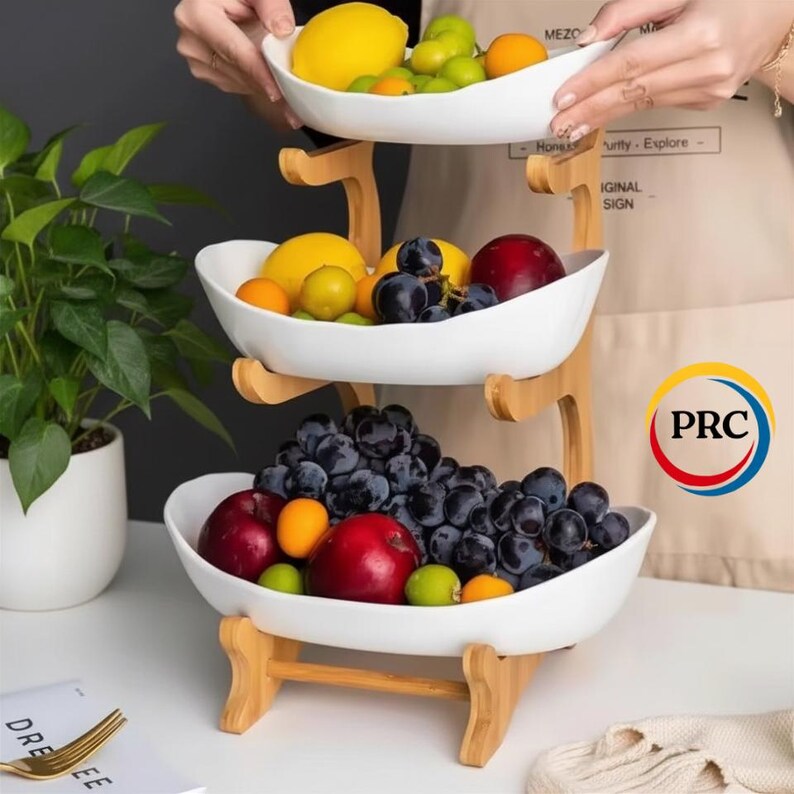 May include: A three-tiered fruit stand featuring white ceramic bowls supported by a light-colored wooden frame. The bowls are filled with assorted fresh fruit, including red apples, dark purple grapes, yellow lemons, and small orange citrus fruits.