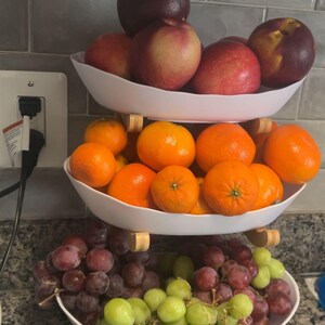 May include: A three-tiered white serving stand featuring three oval bowls stacked vertically. The top bowl holds red and dark purple apples. The middle bowl contains bright orange tangerines. The bottom bowl holds clusters of red and green grapes.