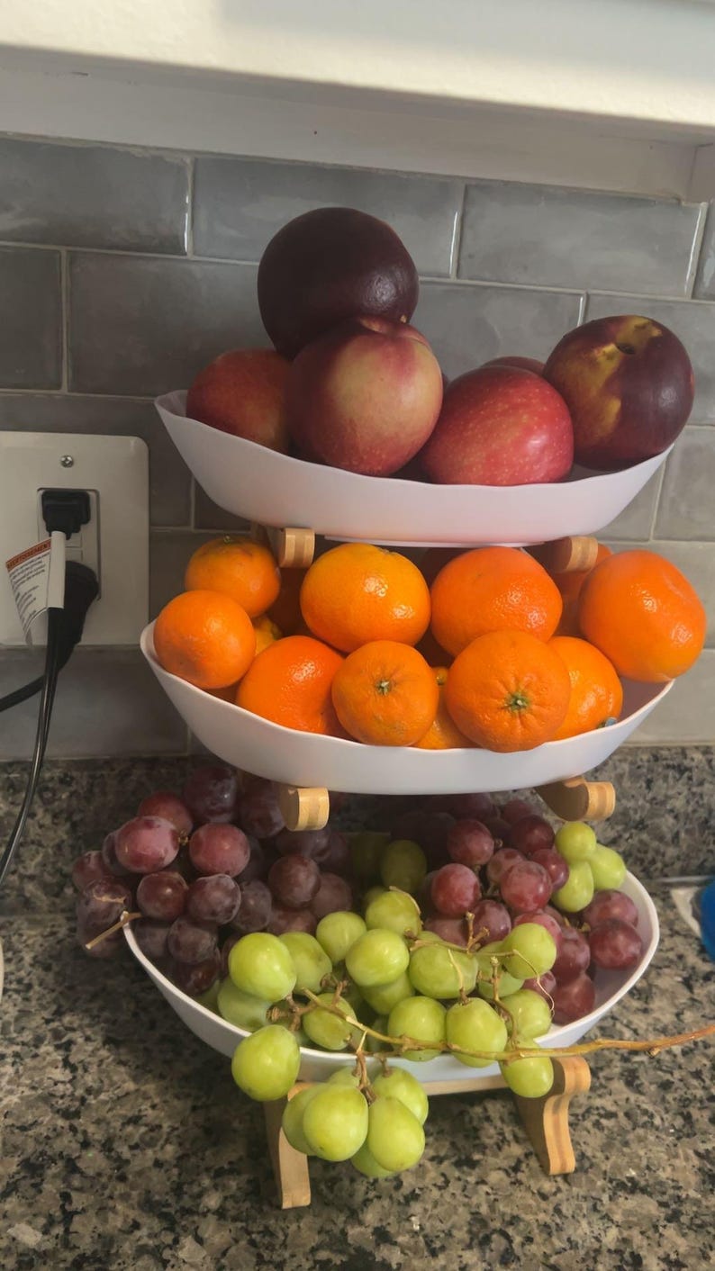 May include: A three-tiered white serving stand featuring three oval bowls stacked vertically. The top bowl holds red and dark purple apples. The middle bowl contains bright orange tangerines. The bottom bowl holds clusters of red and green grapes.