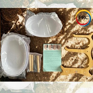 May include: Two white oval bowls wrapped in clear plastic, a light-colored wooden tiered stand, a small plastic bag containing four wooden dowels and a metal tool, and a printed instruction sheet with diagrams on a textured brown surface.