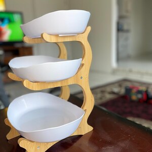 May include: A three-tiered serving stand featuring a light-colored bamboo frame with curved supports. The stand holds three white, oval-shaped bowls with smooth, slightly irregular edges, arranged vertically to provide multiple levels for food display.