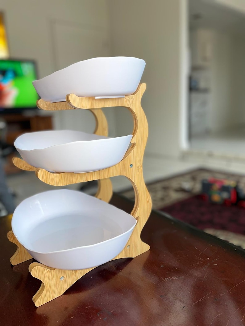May include: A three-tiered serving stand featuring a light-colored bamboo frame with curved supports. The stand holds three white, oval-shaped bowls with smooth, slightly irregular edges, arranged vertically to provide multiple levels for food display.