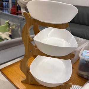 May include: A three-tiered serving stand featuring a light brown bamboo frame with a natural wood grain finish. The stand holds three white, square-shaped ceramic bowls with rounded corners, stacked vertically in a cascading arrangement.