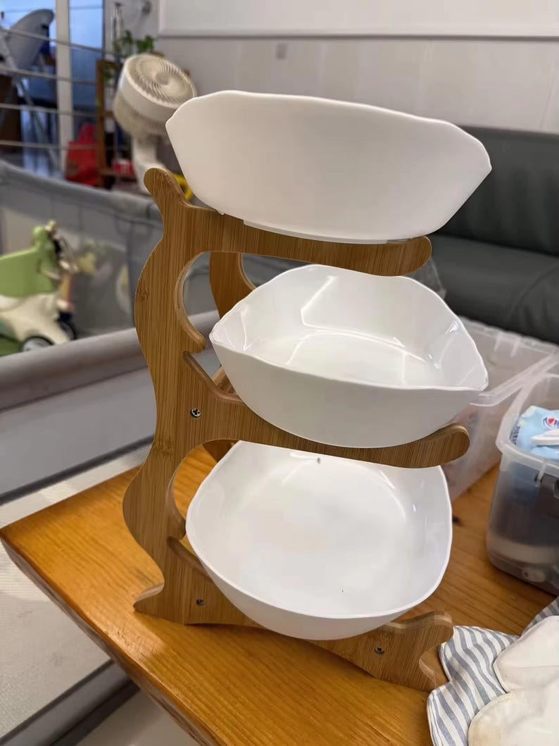 May include: A three-tiered serving stand featuring a light brown bamboo frame with a natural wood grain finish. The stand holds three white, square-shaped ceramic bowls with rounded corners, stacked vertically in a cascading arrangement.