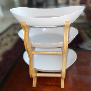 May include: A three-tiered serving stand featuring three white oval-shaped bowls stacked vertically. The bowls are supported by a light-colored wooden frame with curved side supports and horizontal crossbars connecting the structure at the base.
