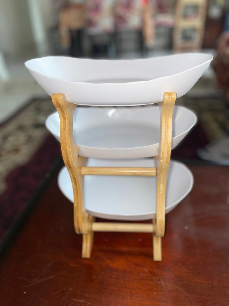 May include: A three-tiered serving stand featuring three white oval-shaped bowls stacked vertically. The bowls are supported by a light-colored wooden frame with curved side supports and horizontal crossbars connecting the structure at the base.