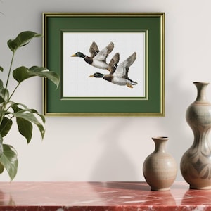 Könnte beinhalten: Gerahmtes Kreuzstich-Kunstwerk mit drei fliegenden Enten. Die Enten sind mit detaillierten Flügeln und Gefieder dargestellt, vor einem weißen Hintergrund. Das Kunstwerk ist in einem grünen Passepartout und einem goldfarbenen Rahmen gerahmt.