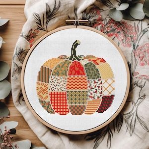 Puede incluir: Calabaza de punto de cruz en un bastidor de bordado de madera. La calabaza está compuesta por varias secciones de estilo patchwork en tonos naranja, rojo, verde y amarillo. El fondo es de tela blanca, y el bastidor descansa sobre una tela con estampado floral.
