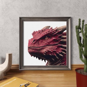 Puede incluir: Arte en punto de cruz enmarcado que representa una cabeza de dragón roja detallada con escamas y cuernos texturizados. La obra de arte se presenta en un marco gris oscuro sobre un fondo blanco. Un cactus en una maceta roja y un libro con una cubierta amarilla están en primer plano.