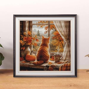 Puede incluir: Una obra de punto de cruz enmarcada que representa a un gato atigrado naranja sentado en el alféizar de una ventana mirando un paisaje otoñal. La escena incluye un jarrón con hojas de otoño, una pequeña calabaza naranja y una taza de café humeante.