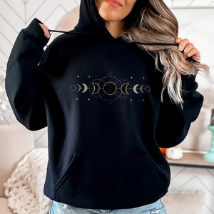 Puede incluir: Sudadera con capucha negra con un dise&ntilde;o de fases lunares doradas. El dise&ntilde;o presenta una luna creciente, luna llena y sol con estrellas.