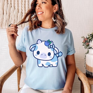 以下が含まれることがあります： ブルーのTシャツに、ブルーベリーのヘッドバンドをした漫画の牛が描かれています。牛は白地に青い斑点があり、かわいい表情をしています。